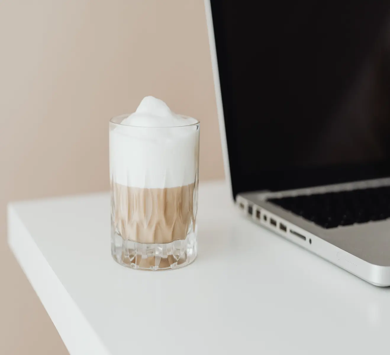 Ein Glas mit Milchschaumgetränk steht neben einem aufgeklappten Laptop auf einem weißen Tisch.