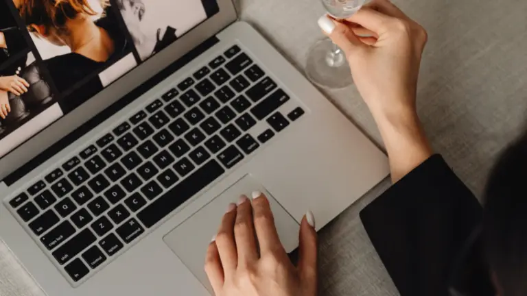 Aus der Vogelperspektive fotografiert: Eine Frau im eleganten schwarzen Blazer arbeitet an einem silbernen Laptop, während sie mit der anderen Hand ein Sektglas hält.