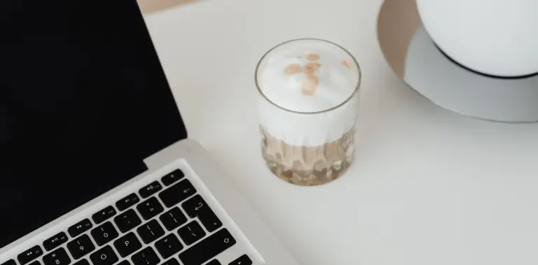 Ausschnitt eines Laptops neben einem Glas Latte Macchiato auf einem weißen Tisch.