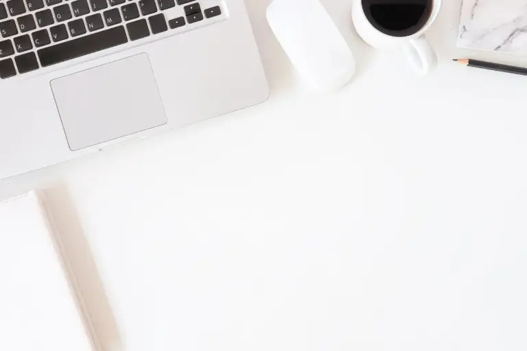 Laptop, Maus, Kaffeetasse und Notizbuch auf weißem Schreibtisch aus der Vogelperspektive