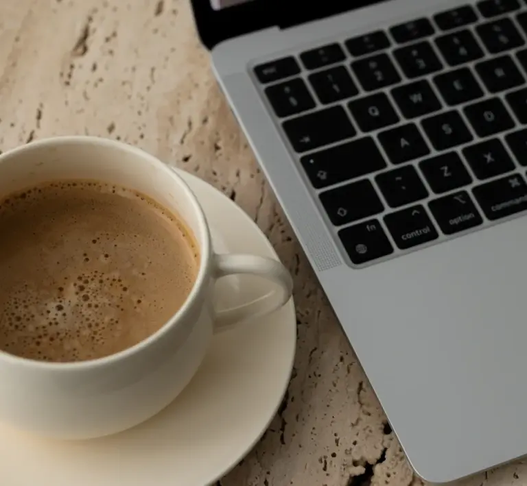 Weiße Tasse mit Kaffee auf Untertasse neben geöffnetem silbernem Laptop auf hellem Tisch