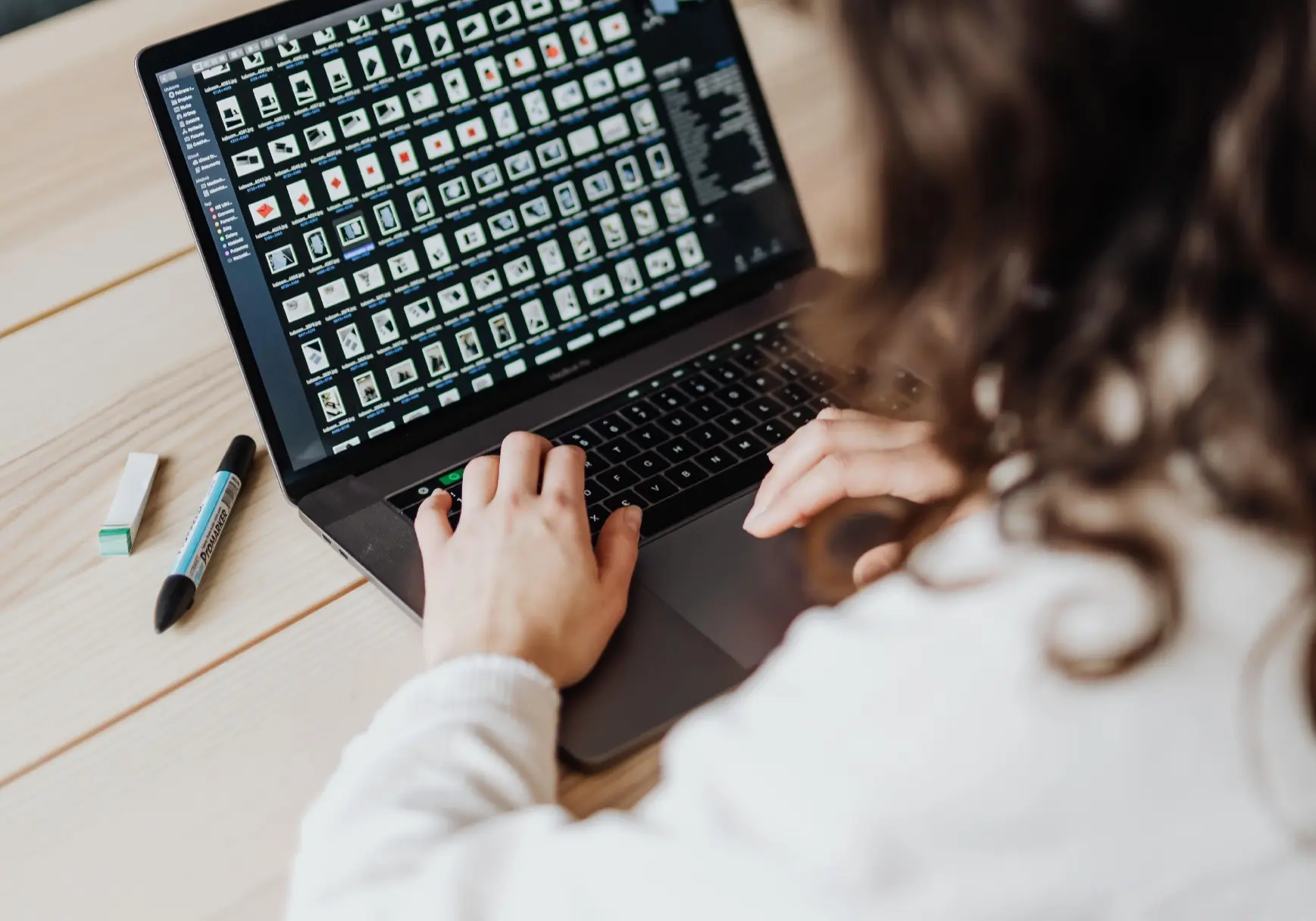 Person mit lockigen Haaren arbeitet an einem Laptop mit geöffneter Bildübersicht, daneben liegen ein Marker und ein Radiergummi auf dem Holztisch.