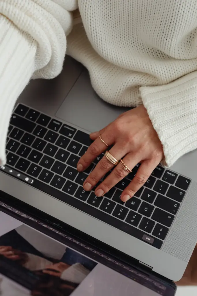 Hand mit mehreren Ringen auf Tastatur eines Laptops, Person trägt grob gestrickten weißen Pullover