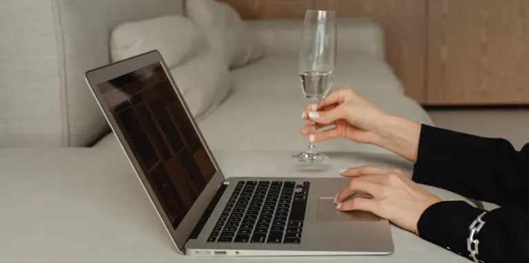 Hände einer Person bedienen ein Laptop auf einem Tisch, die rechte Hand hält ein Glas Sekt, im Hintergrund ein Sofa und ein Schrank.