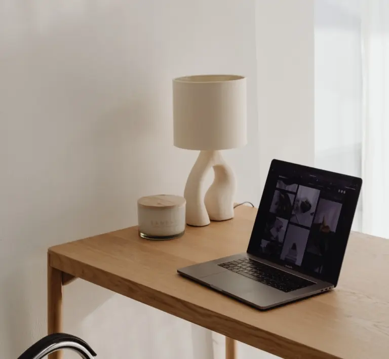Moderner Holztisch mit Laptop, abstrakter weißer Tischlampe und Duftkerze vor weißer Wand und hellem Vorhang